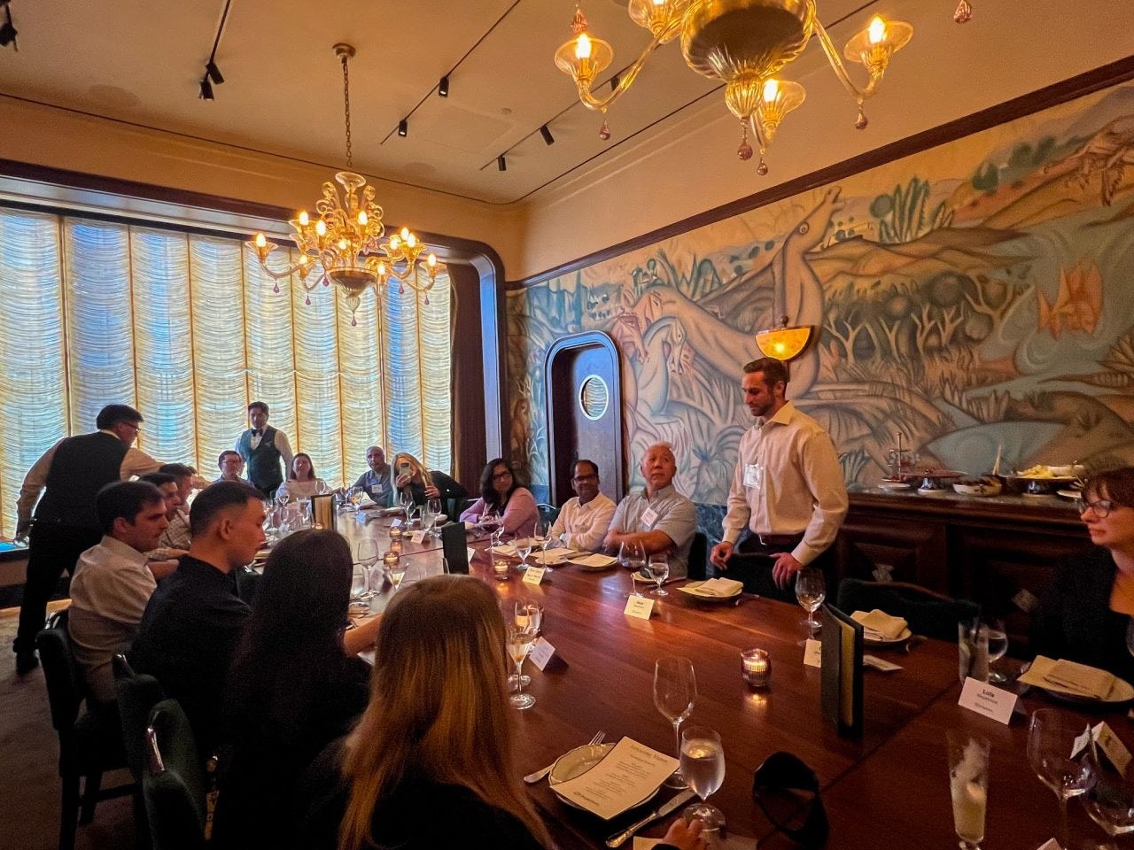 A host standing at the head of a long dinner table addressing seated guests.