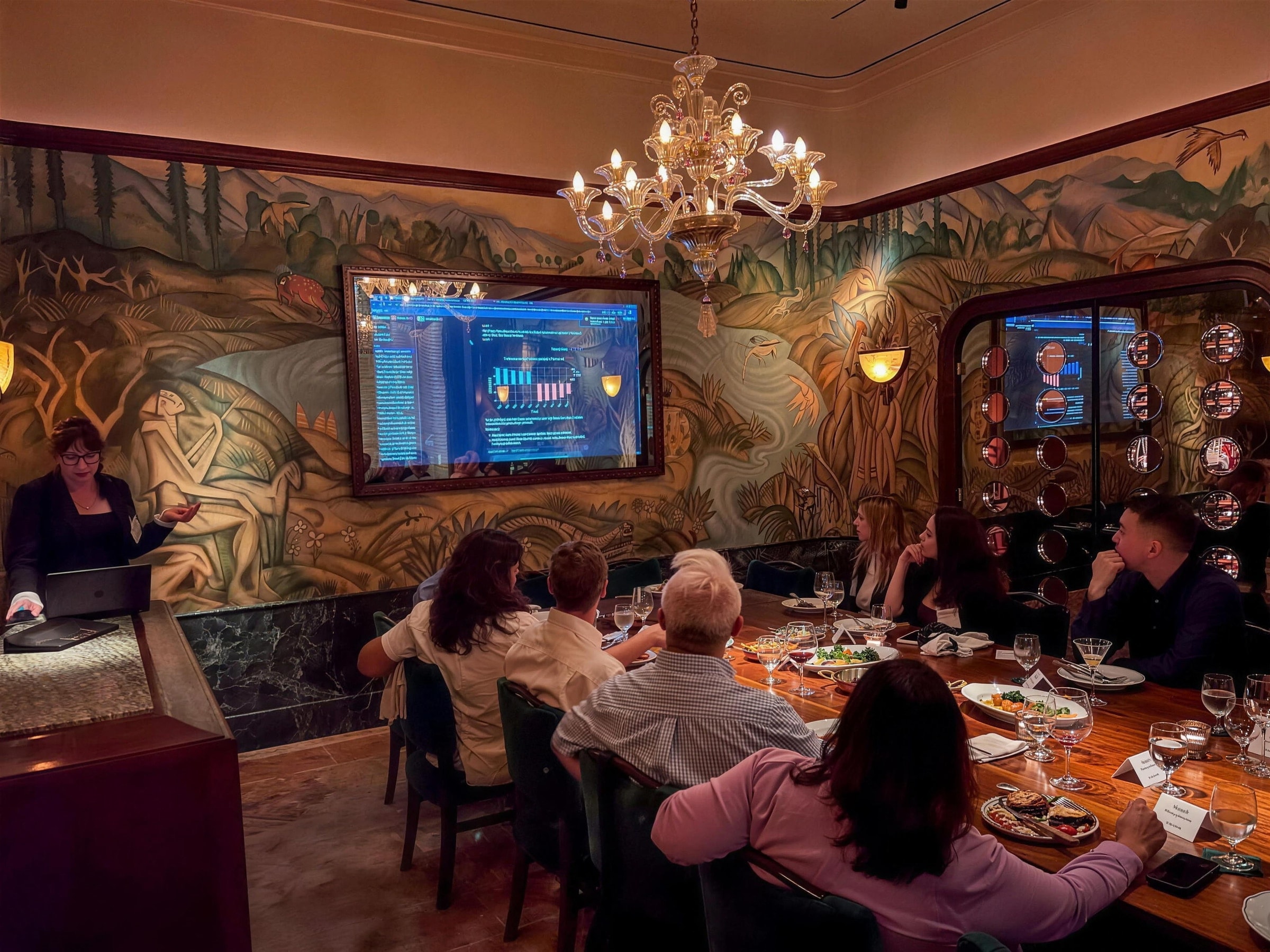 Guests seated around a long table listening to a speaker beside a presentation screen.