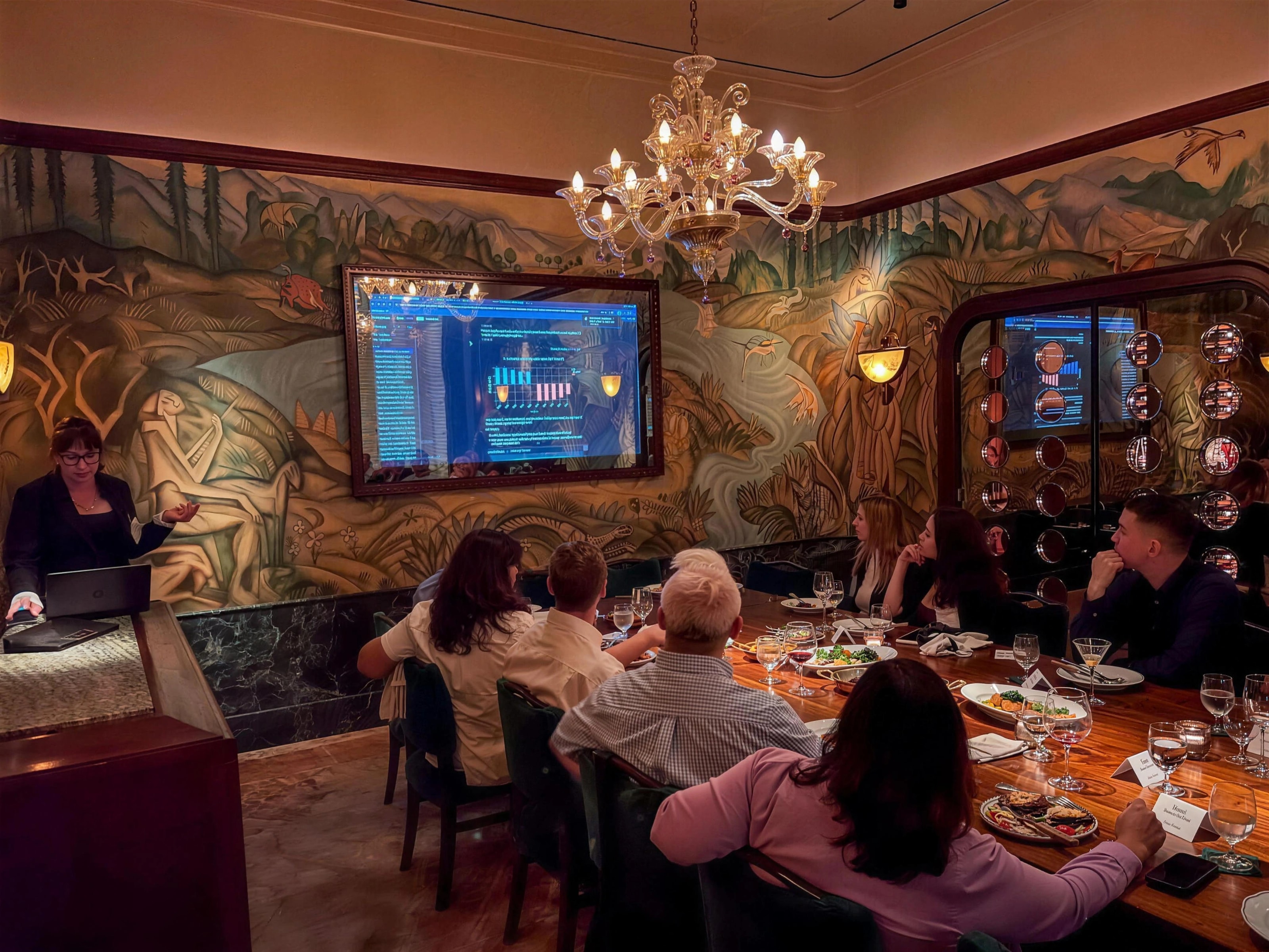 Guests seated around a long table listening to a speaker beside a presentation screen.
