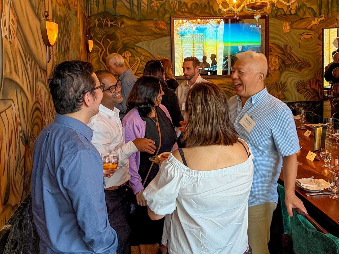 Guests greeting each other with drinks in hand before a dinner, several laughing.